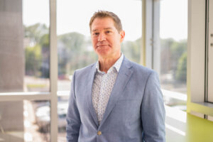 Male in blazer and collared shirt looks at camera standing in front of bright lit windows in a modern office setting.